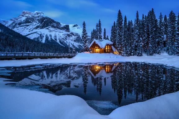 Snowy cabin at dusk by a reflective lake with mountains and pine trees.