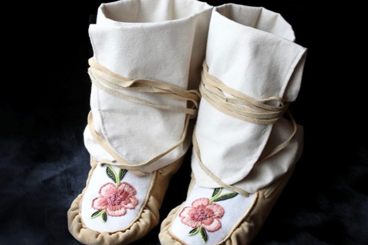 Handmade beige moccasins with floral embroidery on toes, set against a dark background.