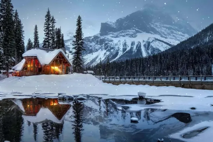 Snowy cabin by a frozen lake with lit windows, surrounded by trees and mountains.