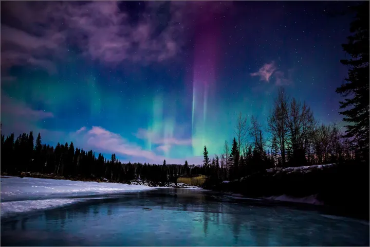 Northern lights over a snowy landscape with trees and a frozen river at night.
