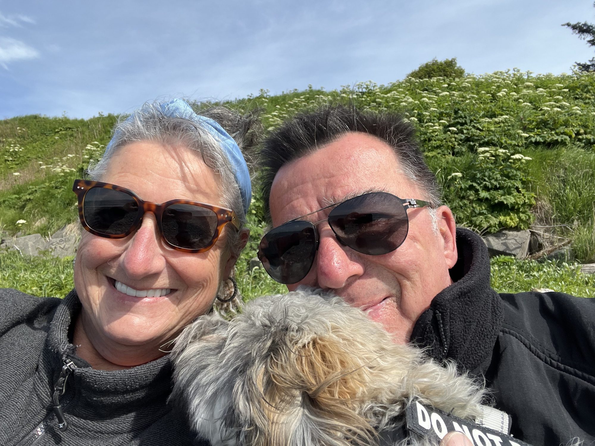 IMG_6697 Two people wearing sunglasses smiling with a dog, green hill in background.