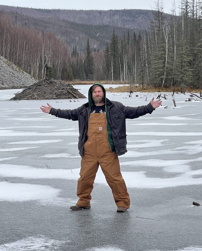 Person in winter clothes with arms outstretched stands on a frozen lake surrounded by trees.