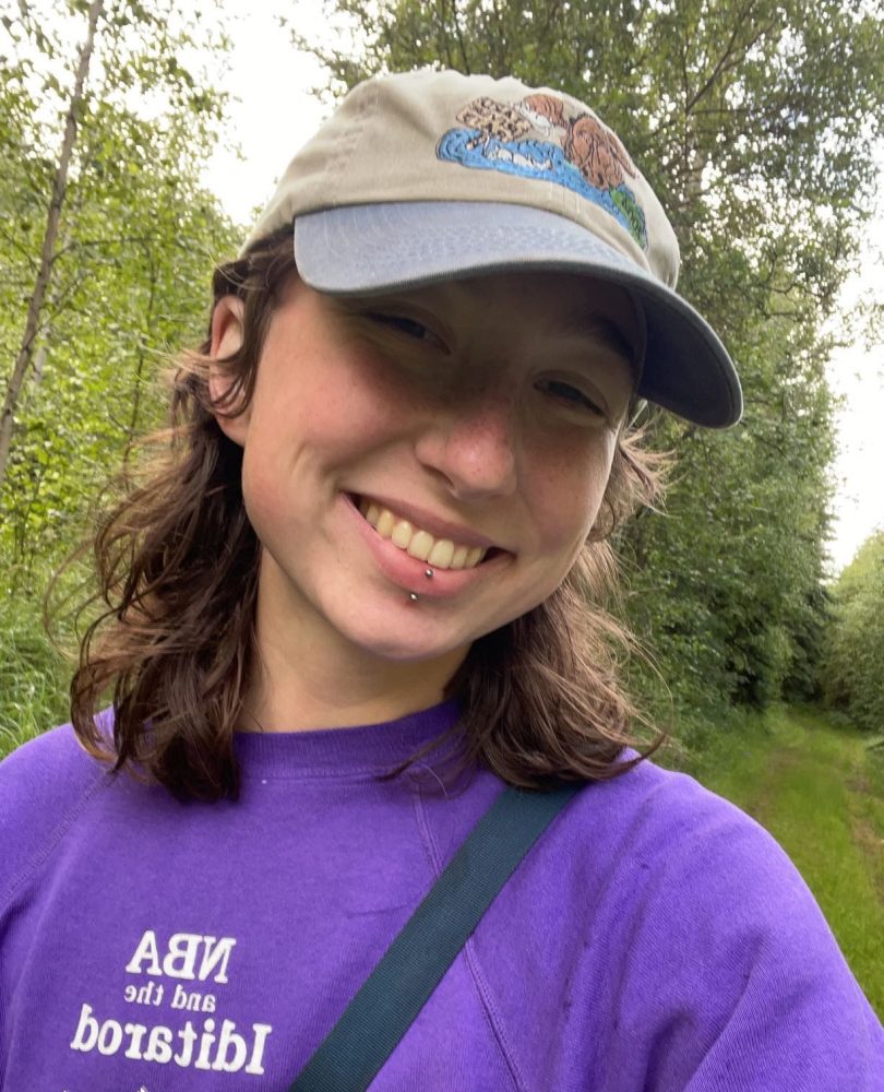 Smiling person in purple shirt and cap on a forest path.