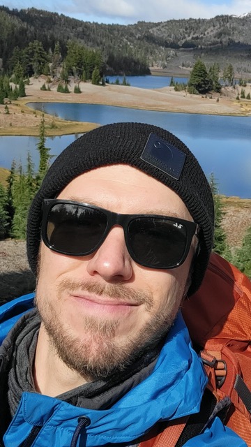 Person in sunglasses and beanie outdoors with a lake and mountains in the background.